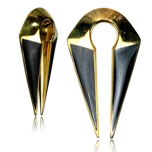 May include: Two gold-colored, geometric ear gauges with a black inlay. The gauges have a teardrop shape with a circular opening at the top. The design is symmetrical, with a pointed bottom and a split center.