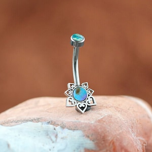 May include: A silver body jewelry barbell with a decorative flower-shaped end featuring a blue and green abalone shell inlay.
