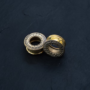 May include: Two gold-colored ear gauges with intricate designs. The gauges are on a black surface.