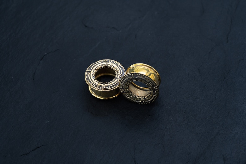 May include: Two gold-colored ear gauges with intricate designs. The gauges are on a black surface.
