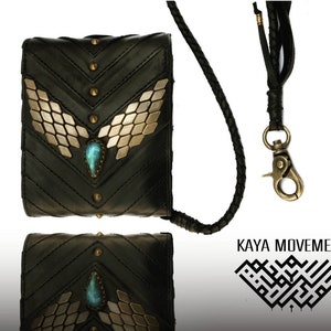 Pode incluir: Uma carteira de couro preta com uma corrente dourada e uma pedra turquesa. A carteira tem um design decorativo de asas douradas e tachas. O nome da marca "KAYA MOVEMENT" est&aacute; impresso em preto sobre um fundo branco.