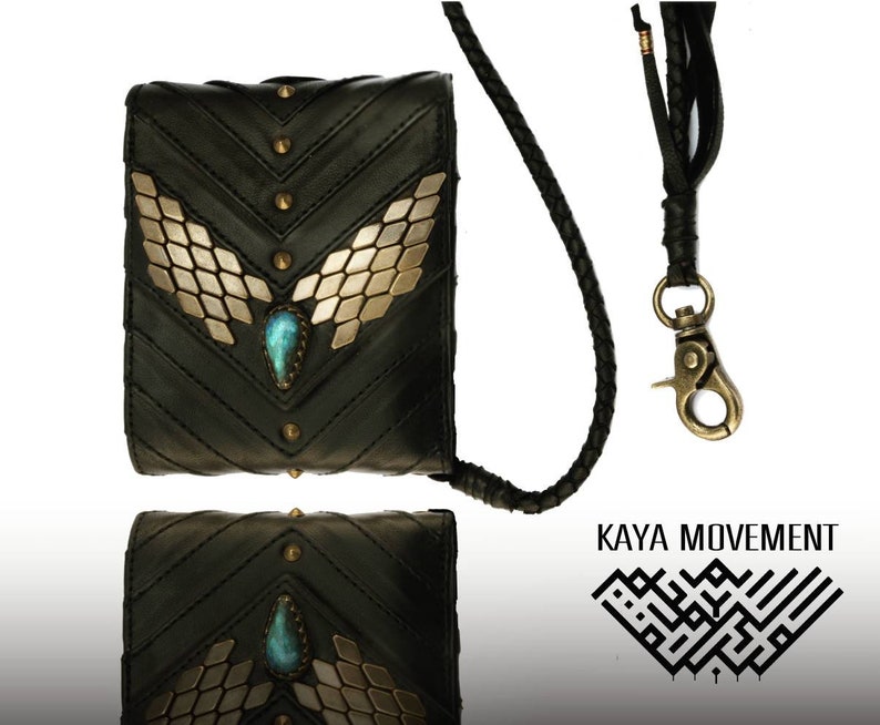 Pode incluir: Uma carteira de couro preta com uma corrente dourada e uma pedra turquesa. A carteira tem um design decorativo de asas douradas e tachas. O nome da marca "KAYA MOVEMENT" est&aacute; impresso em preto sobre um fundo branco.
