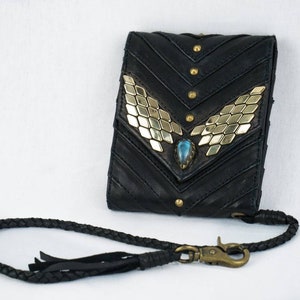 Pode incluir: Uma carteira de couro preta com um detalhe decorativo dourado e azul. A carteira tem uma al&ccedil;a de couro tran&ccedil;ada com um fecho de metal.