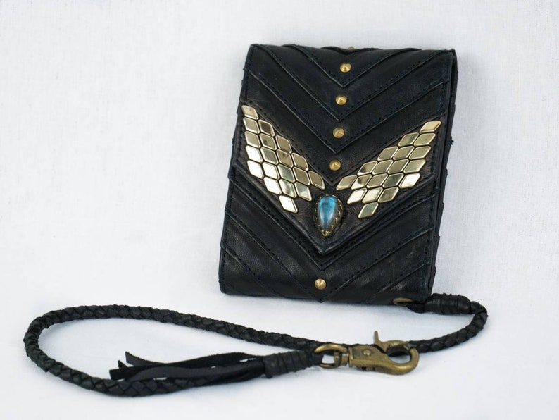 Pode incluir: Uma carteira de couro preta com um detalhe decorativo dourado e azul. A carteira tem uma al&ccedil;a de couro tran&ccedil;ada com um fecho de metal.