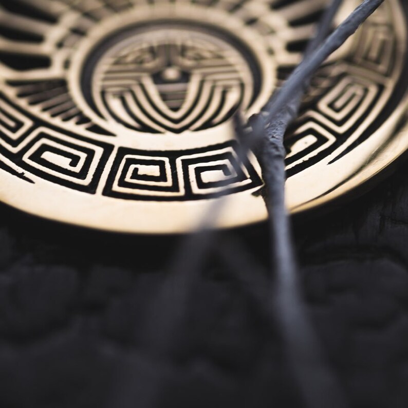 Puede incluir: Un quemador de incienso de metal dorado con un dise&ntilde;o geom&eacute;trico en blanco y negro. Dos varillas de incienso descansan sobre el quemador.
