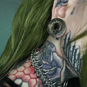 Close-up of a green-haired person with a large gold tunnel in a stretched ear, featuring an alien spaceship design. They have neck tattoos with geometric and floral patterns, and wear a spiked chain necklace, emphasizing a futuristic, cyberpunk style
