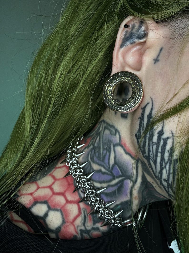 Close-up of a green-haired person with a large gold tunnel in a stretched ear, featuring an alien spaceship design. They have neck tattoos with geometric and floral patterns, and wear a spiked chain necklace, emphasizing a futuristic, cyberpunk style