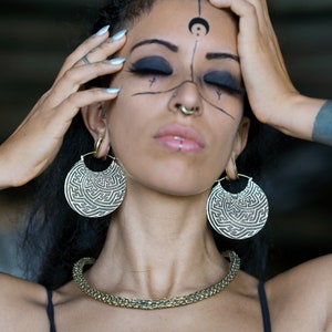 May include: A woman with dark hair and dark eye makeup is wearing large, round, gold earrings with intricate designs. She has a nose ring and a lip piercing. She is wearing a black tank top and a gold chain necklace.
