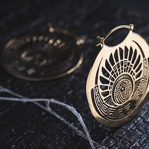 Puede incluir: Un par de pendientes de aro dorados con un dise&ntilde;o geom&eacute;trico. Los pendientes est&aacute;n hechos de metal y tienen una forma circular. El dise&ntilde;o presenta un sol estilizado con rayos que emanan del centro.