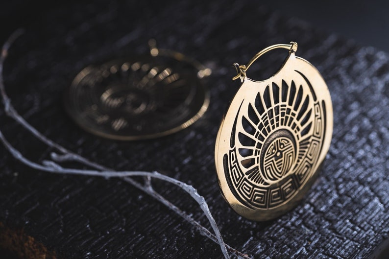 Puede incluir: Un par de pendientes de aro dorados con un dise&ntilde;o geom&eacute;trico. Los pendientes est&aacute;n hechos de metal y tienen una forma circular. El dise&ntilde;o presenta un sol estilizado con rayos que emanan del centro.