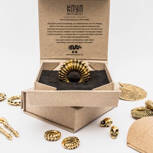 May include: A gold-colored ring with a ribbed design is displayed in an open cardboard box. Several other gold-toned jewelry pieces, including rings, pendants, and a skull-shaped charm, are scattered around the box. The box has text on the inside.