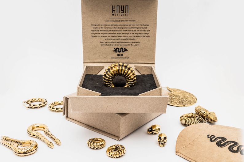 May include: A gold-colored ring with a ribbed design is displayed in an open cardboard box. Several other gold-toned jewelry pieces, including rings, pendants, and a skull-shaped charm, are scattered around the box. The box has text on the inside.