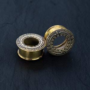 May include: Two gold-colored ear gauges with intricate designs. The gauges are on a black surface.