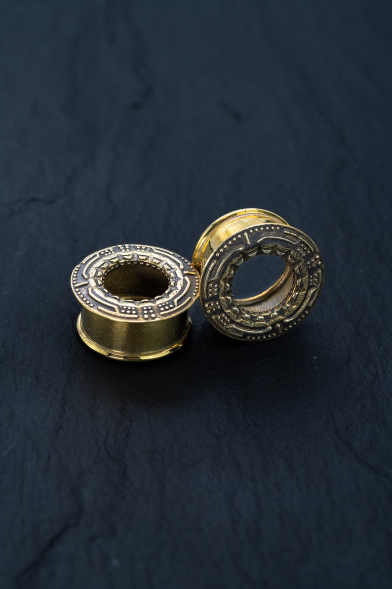 May include: Two gold-colored ear gauges with intricate designs. The gauges are on a black surface.