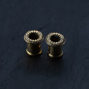 May include: Two gold-colored ear gauges with intricate designs around the edges. The gauges are on a dark gray surface.