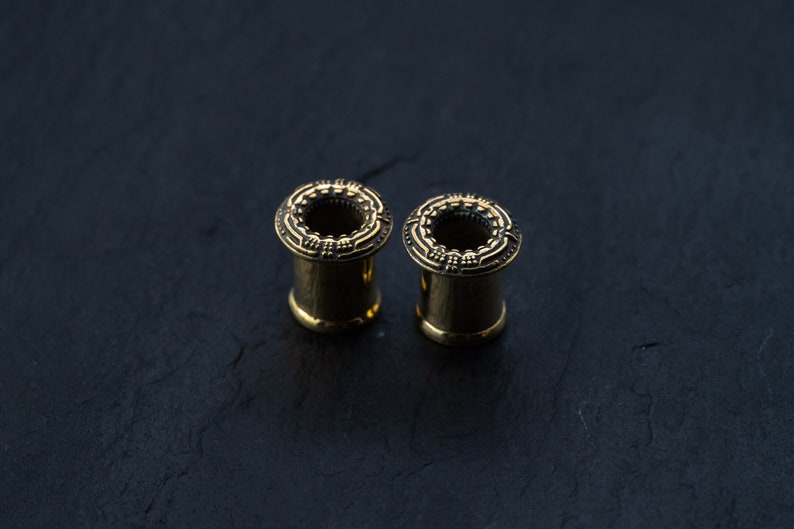 May include: Two gold-colored ear gauges with intricate designs around the edges. The gauges are on a dark gray surface.