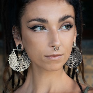 May include: A woman with dark hair and a silver nose ring is wearing large silver earrings with a honeycomb design. She is also wearing a silver hoop earring in her left ear.