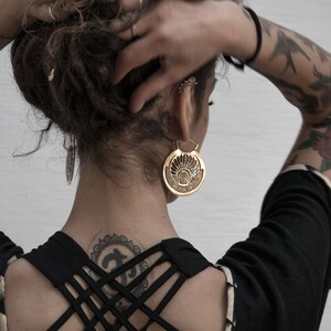 Puede incluir: Una mujer con el pelo oscuro y tatuajes lleva una blusa negra con la espalda cruzada. Lleva un pendiente grande de oro adornado en una oreja.