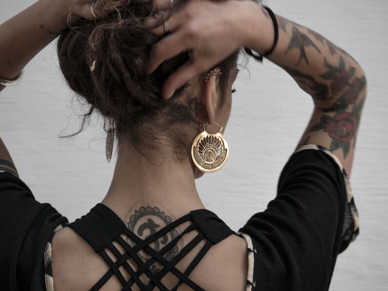 Puede incluir: Una mujer con el pelo oscuro y tatuajes lleva una blusa negra con la espalda cruzada. Lleva un pendiente grande de oro adornado en una oreja.