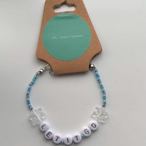 Könnte beinhalten: Ein blaues und weißes Perlenarmband mit weißen Buchstabenperlen, die "Let It Go" buchstabieren, und Schneeflockenanhängern. Das Armband befindet sich auf einer braunen Karte mit dem Text "Emily's Version".