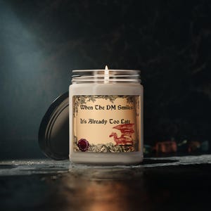 May include: A white candle with a gold and red label that reads "When the DM Smiles It's Already Too Late". The label features a red dragon with a gold background.