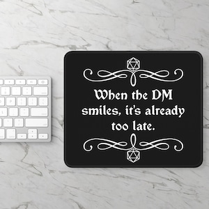 May include: Black mousepad with white text that reads "When the DM smiles, it's already too late."  The text is framed by two decorative swirls and a 20-sided die at the top and bottom.