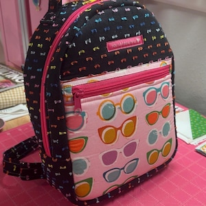 May include: A black backpack with a textured surface featuring small colorful dashes. The front pocket displays a pink fabric with a pattern of various colored eyeglasses. A small rectangular label on the top front reads YOU ARE BEAUTIFUL.