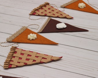 Könnte beinhalten: Ein festliches Banner mit wimpelförmigen Tortenstücken. Einige Wimpel sind orange, braun und beige, mit weißen Blumenakzenten. Die Tortenboden-Wimpel haben ein gewebtes Design. Jeder Wimpel ist an einer natürlichen Schnur aufgereiht.