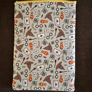 May include: A light blue book cover featuring a repeating pattern of brown sorting hats, gold glasses, red and gold ties, white envelopes, and gold wands. The cover is edged with yellow fabric.