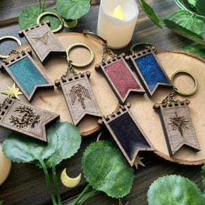 May include: A collection of wooden keyrings in a flag design, featuring engraved patterns and coloured resin. The keyrings are displayed on a wooden slice, with decorative leaves and a candle, and have bronze-toned metal fixings.