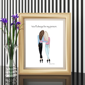 K&ouml;nnte beinhalten: Ein gerahmter Druck in einem goldfarbenen Rahmen zeigt eine Illustration von zwei Figuren, die sich umarmen. Die Figuren tragen Jeans und Oberteile in Blau und Rosa. Der Text "You'll always be my person" steht &uuml;ber den Figuren.