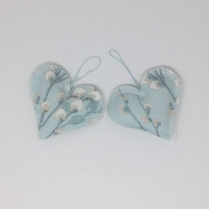 May include: Two heart-shaped decorations with a pale blue fabric and a floral design in white and grey. Each has a loop for hanging. The edges are stitched with a contrasting thread.