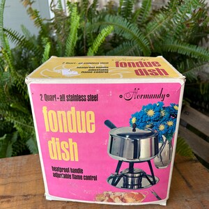 May include: A vintage "Normandy" fondue dish box with a pink and yellow design. The box features a stainless steel fondue pot illustration, with text indicating a 2-quart capacity, heatproof handle, and adjustable flame control.