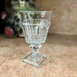 May include: A clear, vintage-style glass goblet with a square base and a detailed, textured design. The goblet has a short stem and a wide, fluted bowl. A panda bear cookie jar is in the background.