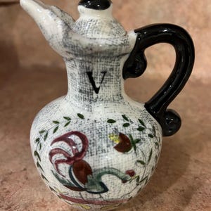 May include: A small, ceramic pitcher with a black handle and spout. The pitcher is white with a gray textured pattern and a rooster illustration. The letter "V" is printed on the front.