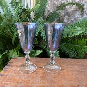 May include: Two elegant, stemmed wine glasses with a smoky, iridescent finish. The bowls are a deep purple-blue, transitioning to clear stems with decorative ball accents. The glasses sit on a wooden surface, with green foliage in the background.