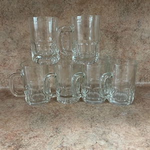 May include: A set of seven clear glass beer mugs with handles. The mugs have a classic, paneled design and are arranged on a brown surface. The mugs are empty and ready to be filled.