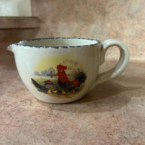May include: Cream-colored ceramic creamer with a handle and spout. The creamer features a rooster and hen illustration in a pastoral scene. The rim has a blue dotted pattern. The creamer is designed for serving cream or milk.