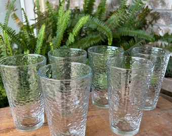 May include: Six clear drinking glasses with a textured, dimpled surface. The glasses are cylindrical and sit on a wooden surface, with green ferns in the background. The glasses are empty and appear to be made of glass.