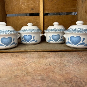 May include: Four white ceramic lidded bowls with a blue heart design. The bowls have small handles and a decorative blue pattern around the rim. The bowls are arranged on a wooden surface.