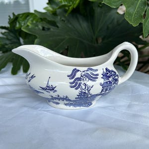 May include: A white ceramic gravy boat with a blue Willow pattern design. The boat features a spout, handle, and a detailed landscape scene. The gravy boat is a classic piece of tableware.