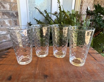 May include: Four clear drinking glasses with a textured, bubble-like pattern. The glasses are tall and cylindrical, perfect for water, juice, or other beverages. They are arranged on a weathered wooden surface, with greenery in the background.