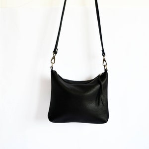 May include: Black leather crossbody bag with a textured finish. The bag features a zipper closure, a long adjustable strap, and brass-colored hardware. The bag is hanging against a white background.