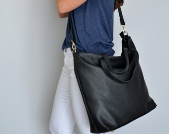 May include: A black leather tote bag with a long strap worn over the shoulder. The bag has a top zipper closure and two top handles. The person wearing the bag is wearing white jeans and a blue shirt.