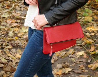 May include: A red leather crossbody bag with a flap closure and a zipper detail. The bag is worn over a person's shoulder and is hanging down the side of their body.