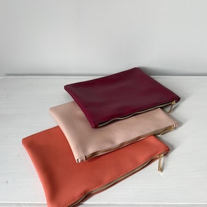 May include: Three rectangular zipped pouches stacked on top of each other. The top pouch is deep burgundy, the middle pouch is a soft pale pink, and the bottom pouch is a vibrant coral orange. Each pouch features a gold-toned metal zip fastener.