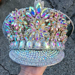 May include: A silver and white hat with iridescent rhinestones and a large flower design. The hat is decorated with a variety of sizes and shapes of rhinestones.