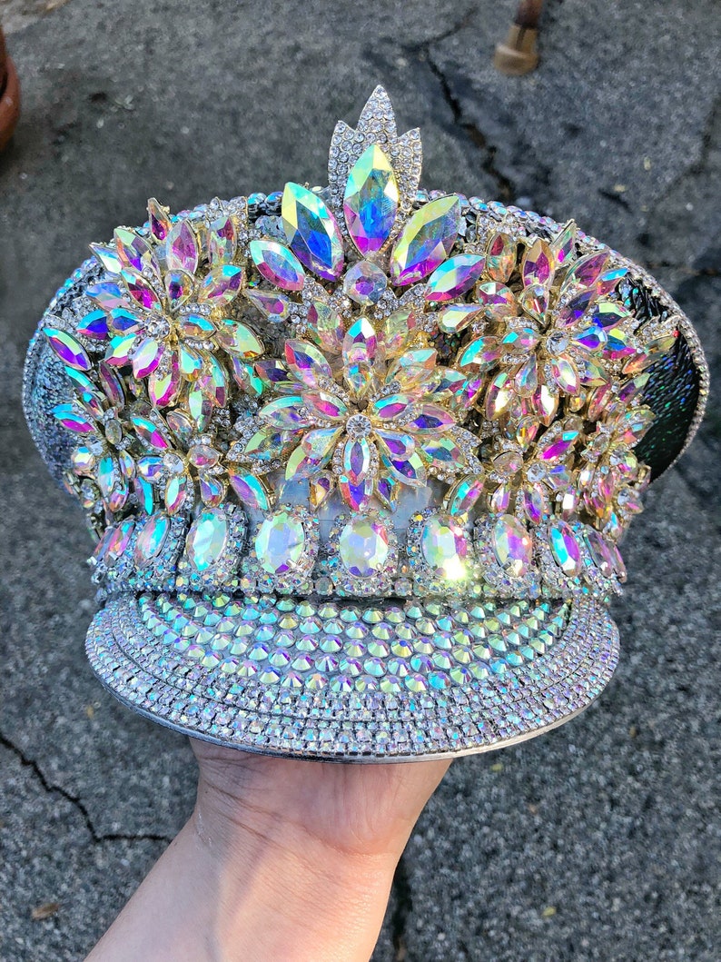 May include: A silver and white hat with iridescent rhinestones and a large flower design. The hat is decorated with a variety of sizes and shapes of rhinestones.