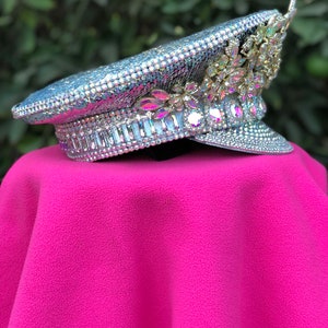 May include: A silver sequined and crystal embellished hat with a gold floral design. The hat is on a pink surface.