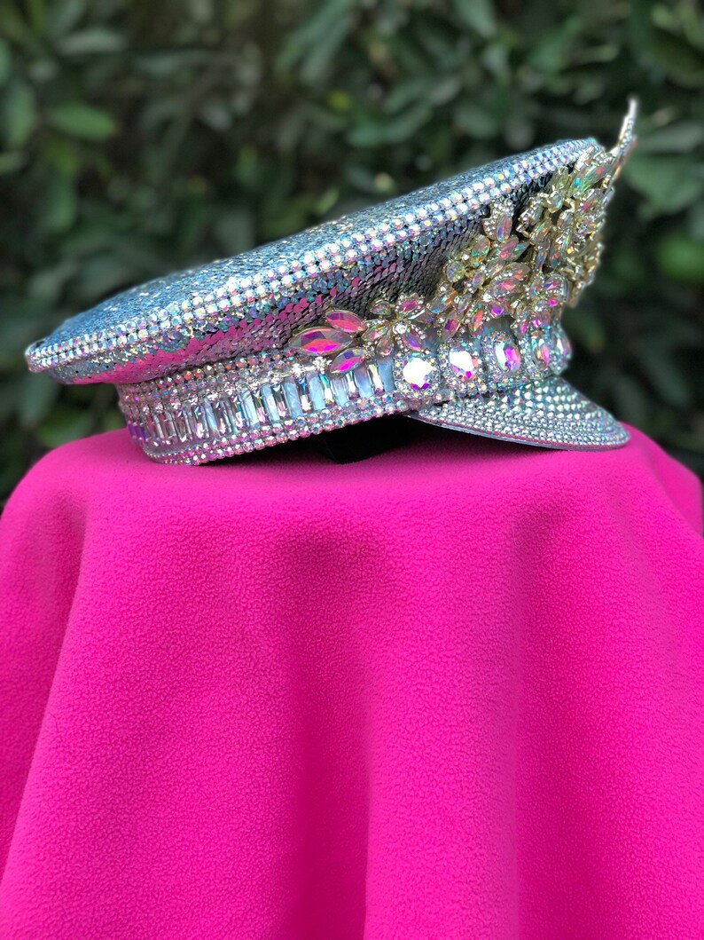 May include: A silver sequined and crystal embellished hat with a gold floral design. The hat is on a pink surface.
